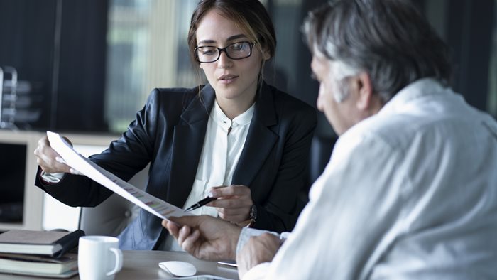 A woman in a business suit sitting behind a desk shows a document on enabling cost transparency in law practice to a male client.